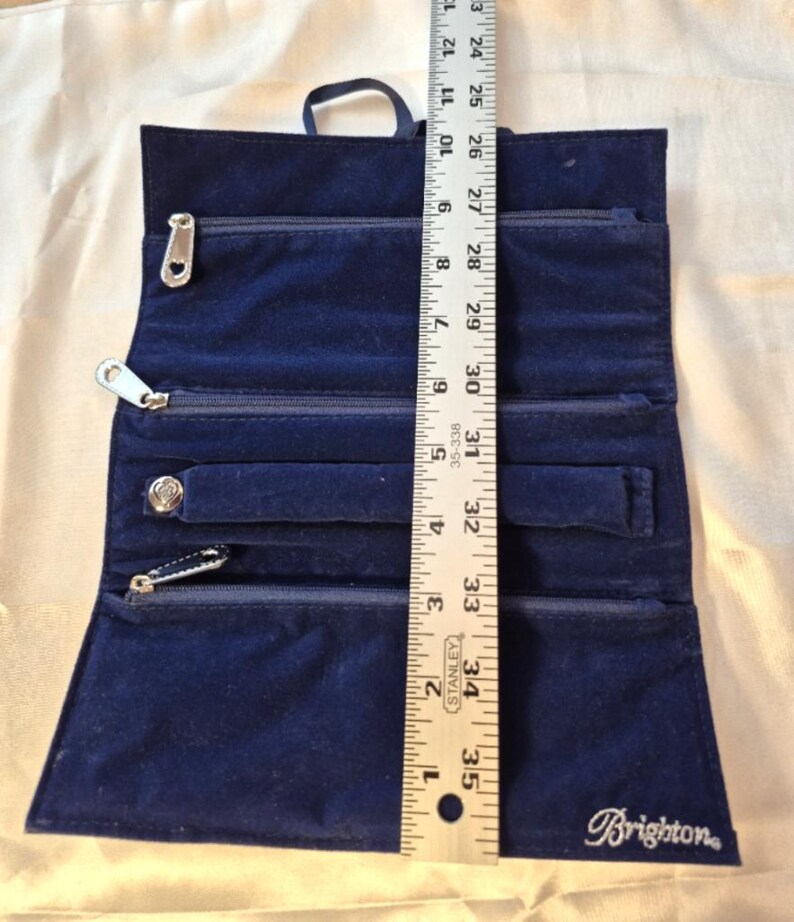 Peut inclure: Un &eacute;tui &agrave; bijoux en velours bleu fonc&eacute;, ouvert pour afficher plusieurs compartiments zipp&eacute;s et une fermeture &agrave; bouton-pression. Le rouleau mesure environ 30 cm de hauteur. Le logo Brighton est visible.