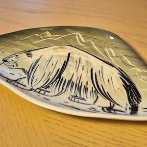 May include: A triangular ceramic dish featuring a stylized black ink illustration of a polar bear in profile. The background is a muted sage green with faint white line drawings of mountain peaks. The dish has a smooth, glossy finish.