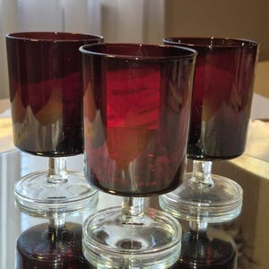 May include: Three ruby red wine glasses with a clear glass base and stem. The glasses have a cylindrical shape and are displayed on a reflective surface. The glasses are empty and appear to be made of glass.