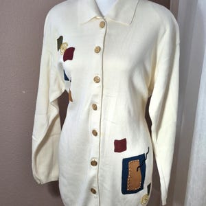 May include: A long-sleeved cream-colored button-down cardigan featuring a collar and a series of gold-toned buttons. The garment is decorated with various geometric patches in shades of dark red, navy blue, and tan, accented with gold studs.