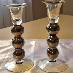May include: Two clear glass candle holders with a brown, spherical design. The candle holders have a flared top and a round base. The spherical design is made of three stacked spheres. The candle holders are a decorative home decor item.