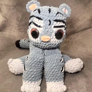 May include: A handmade plush tiger with a light blue body, white paws, and black stripes. The tiger has large, round eyes, a peach-colored nose, and small ears. The plush toy is made of soft, textured yarn.