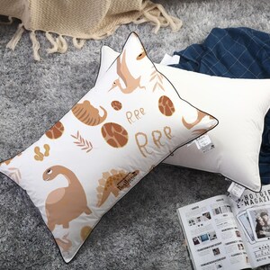 May include: Two rectangular pillows with a white base colour. One pillow features a repeating pattern of brown dinosaurs, eggs, and the text RRR. Both pillows have a thin dark brown piped edge and a small fabric tag attached to the side seam.