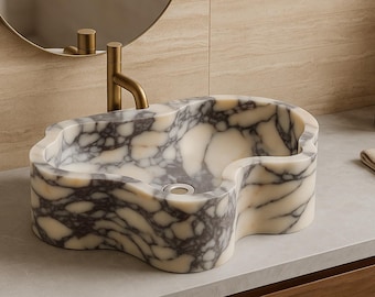 May include: A marble bathroom sink with a distinctive, flowing shape. The sink has a white and grey veined pattern, a drain, and a gold-coloured tap. The sink is set against a neutral-toned wall and countertop.