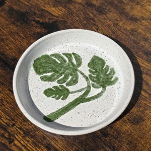 May include: A round, white ceramic dish with a speckled surface. The dish features a hand-painted green monstera leaf design. The rim is slightly raised, and the overall aesthetic is rustic and natural.