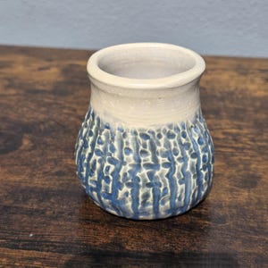 May include: A small, handmade ceramic vase with a textured blue and white exterior. The vase has a cylindrical shape with a slightly flared rim. The base is a dark blue, transitioning to a lighter blue and white pattern. The rim is a creamy white color.