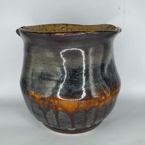 May include: A ceramic pot with a wide, flared rim and a rounded body. The exterior features a dark, mottled glaze in shades of charcoal and black, transitioning into a warm, amber-toned drip pattern near the base. The interior is a light tan.
