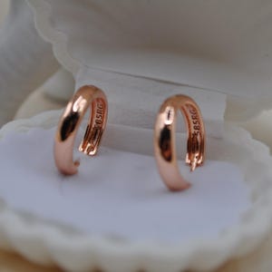 Puede incluir: Un par de pendientes de aro de oro rosa exhibidos en una caja de joyería blanca con forma de concha. Los pendientes son brillantes y tienen un diseño liso y curvado. Cada pendiente tiene la marca "585RG".