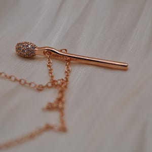 May include: Rose gold matchstick pendant necklace. The match head is encrusted with small, sparkling stones. The chain and matchstick pendant are a warm, metallic color, resting on a soft, white fabric.