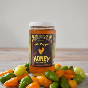 May include: A jar of "Mosquito Lagoon" Datil Pepper Hot Honey sits amongst a pile of colourful peppers. The label features text and illustrations of peppers. The peppers are green, orange, and yellow.