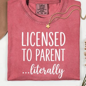 May include: A red-pink t-shirt with the words "LICENSED TO PARENT ...literally" in white. A gold star pendant necklace and a brown hat are also visible. The shirt has a Comfort Colors tag.