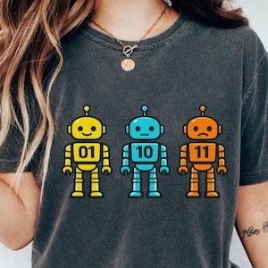 Peut inclure: T-shirt gris foncé avec trois illustrations de robots colorés. Les robots sont jaunes, bleus et oranges, chacun avec un numéro différent : 01, 10 et 11. Le t-shirt est en matière souple.