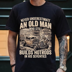 May include: Black t-shirt with a graphic featuring a vintage hot rod and the text "NEVER UNDERESTIMATE AN OLD MAN WHO STILL BUILDS HOTRODS IN HIS SEVENTIES". The design includes a classic car and a mechanic.