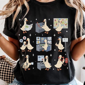 Puede incluir: Camiseta negra con un divertido diseño de gansos de dibujos animados en roles de servicios de emergencia. Los gansos se muestran con periódicos, auriculares, mapas y teléfonos. La camiseta incluye el texto "911 Dispatch".