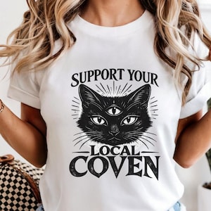 Peut inclure: T-shirt blanc avec une illustration de chat noir à trois yeux et le texte "SUPPORT YOUR LOCAL COVEN". Le motif est imprimé sur le devant du t-shirt. Le t-shirt a un col rond et des manches courtes.