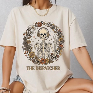 Puede incluir: Camiseta de color marfil con un esqueleto con auriculares y haciendo el signo de la paz, rodeado por una corona floral. El texto "THE DISPATCHER" está debajo. La camiseta es de manga corta y de estilo casual.