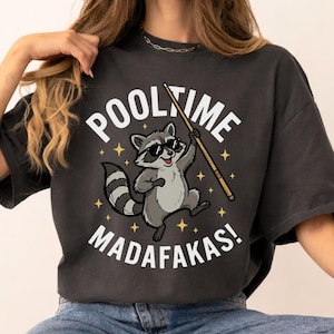 May include: Dark gray t-shirt featuring a cartoon raccoon wearing sunglasses and holding a pool cue. The text "POOL TIME MADAF*KAS!" is printed above and below the raccoon illustration. The shirt is a relaxed fit.