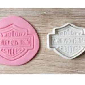 Op de afbeelding: Een roze siliconen Harley-Davidson koekjessnijder naast een witte plastic snijder. De roze snijder heeft het Harley-Davidson logo in reliëf, terwijl de witte snijder het spiegelbeeld laat zien. Beide liggen op een licht houten oppervlak.