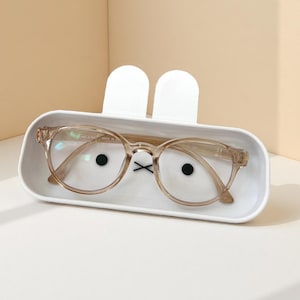 May include: A pair of clear-framed eyeglasses rests inside a white, bunny-shaped glasses case. The case has two white, upright ear-shaped protrusions. The glasses have a round shape and are light brown. Black dots and a black 'X' are printed on the case.