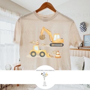May include: A beige t-shirt featuring a construction theme. The design includes yellow construction vehicles, traffic cones, and clouds. The text "WILDFLOWER - HONEY DESIGN CO." is printed at the bottom. The shirt is hanging on a wooden hanger.