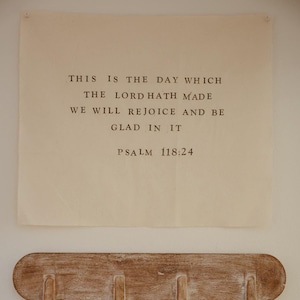 May include: A beige rectangular wall hanging with the text "THIS IS THE DAY WHICH THE LORD HATH MADE WE WILL REJOICE AND BE GLAD IN IT PSALM 118:24" in black font. Below it is a wooden coat rack with four pegs.