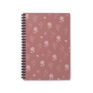 May include: A spiral-bound notebook with a dusty rose cover. The cover features a repeating pattern of delicate, white line-drawn flowers and foliage. The notebook's black spiral binding is visible along the left edge.