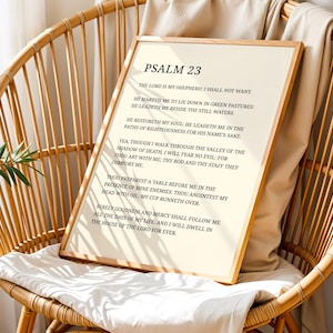 May include: A framed print featuring the text of Psalm 23 in black font on an off-white background. The print has a light wood frame and is propped on a white cushion inside a wicker chair. The chair is made of light brown woven material.