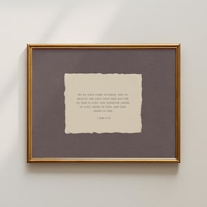 May include: A framed print with a gold-coloured frame. The print features a torn-edge paper with text from 1 John 4:16. The background is a muted purple colour.