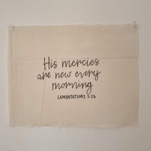 May include: A rectangular, off-white canvas with the text "His mercies are new every morning" and "LAMENTATIONS 3:23" in black script. The canvas is attached to a white wall with two clear push pins.