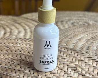 May include: A white serum bottle with a wooden cap and dropper. The bottle has the text "SERUM DE VISAGE SAFRAN" and "30ml" printed on it. The bottle is sitting on a woven surface.
