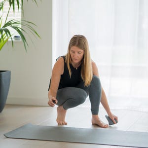 May include: A person wearing a black vest and grey leggings kneels on a grey yoga mat, holding a small black object and a grey massage tool. A large potted plant is visible in the background. The scene is indoors.