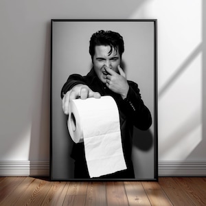 May include: Monochrome framed print showing a person holding out a roll of toilet paper. The person is wearing a dark jacket and has their finger on their nose. The image is set against a neutral background.