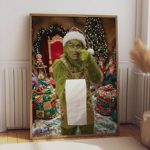 May include: A framed wall art print featuring a green, furry creature wearing a Santa hat and an apron, holding a roll of white toilet paper. The background shows a decorated Christmas tree, a red chair, and gift bags with colorful patterns.