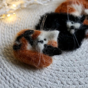 Könnte beinhalten: Drei handgefertigte Katzenfiguren aus Filz in Orange, Schwarz und Weiß. Die Katzen sind zusammengerollt, mit sichtbaren Schnurrhaaren und kleinen schwarzen Perlenaugen. Die Figuren befinden sich auf einer weißen, strukturierten Oberfläche.