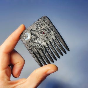 May include: A dark gray, square-shaped hair comb featuring an engraved howling wolf design. The wolf has a small red stone for an eye and is accented by a crescent moon inlay. The surface has a textured, matte finish with leaf motifs.