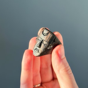 May include: A dark brown wooden ring carved with a stylized face. The face has closed eyes, a small mouth, and a crescent moon carved on the forehead. The ring is held in a hand against a gray background.