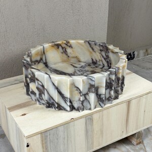 May include: A round, marble sink with a geometric, ridged exterior. The sink features a cream and brown marbled pattern. It is displayed on a light-colored wooden box. The sink's design is modern and would be a statement piece in any bathroom.