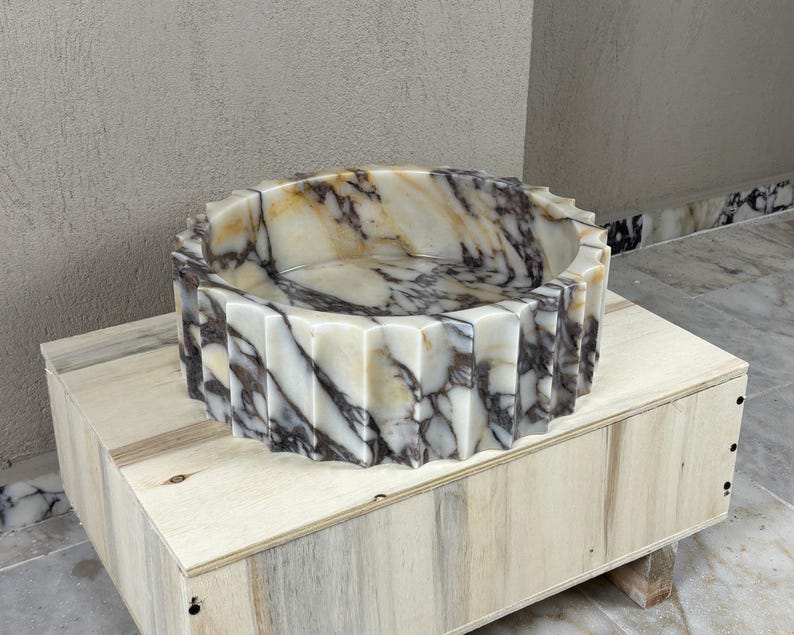 May include: A round, marble sink with a geometric, ridged exterior. The sink features a cream and brown marbled pattern. It is displayed on a light-colored wooden box. The sink's design is modern and would be a statement piece in any bathroom.