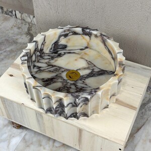 May include: A round marble sink with a scalloped edge. The sink features a cream and gray marbled pattern and a gold-colored drain. It is set on a light-colored wooden box.