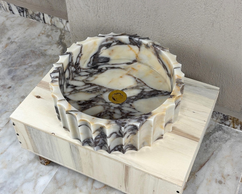 May include: A round marble sink with a scalloped edge. The sink features a cream and gray marbled pattern and a gold-colored drain. It is set on a light-colored wooden box.