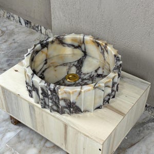 May include: A round, marble sink with a fluted edge, featuring a gold-colored drain. The sink has a cream and gray marbled pattern. It rests on a light-colored wooden cabinet. The sink is a bathroom fixture.