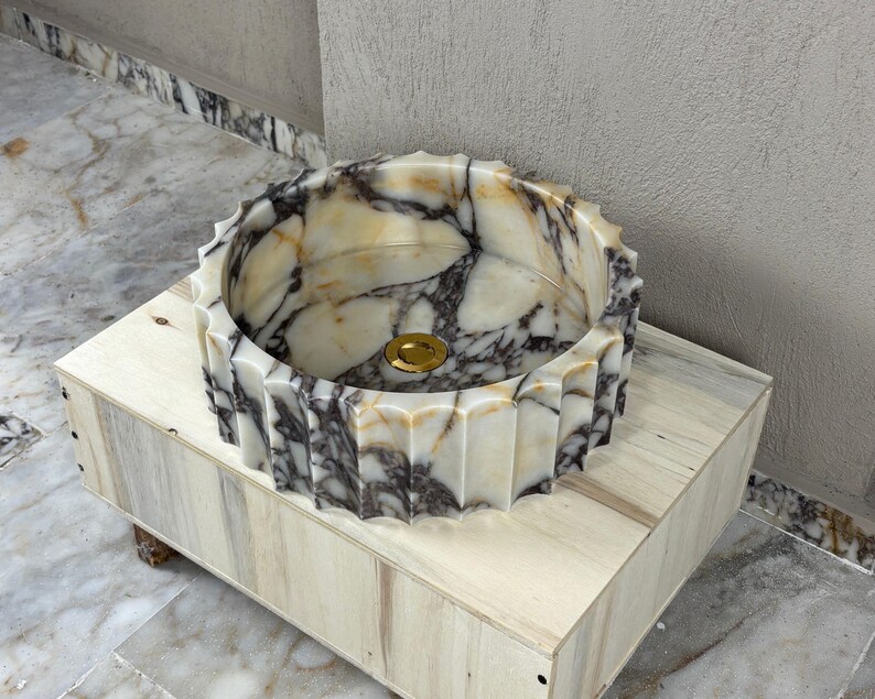 May include: A round, marble sink with a fluted edge, featuring a gold-colored drain. The sink has a cream and gray marbled pattern. It rests on a light-colored wooden cabinet. The sink is a bathroom fixture.