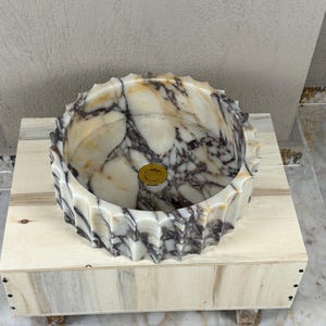 May include: A marble vessel sink with a scalloped edge. The sink features a cream and brown marbled pattern with a gold-colored drain cover. It is set on a light-colored wooden crate.