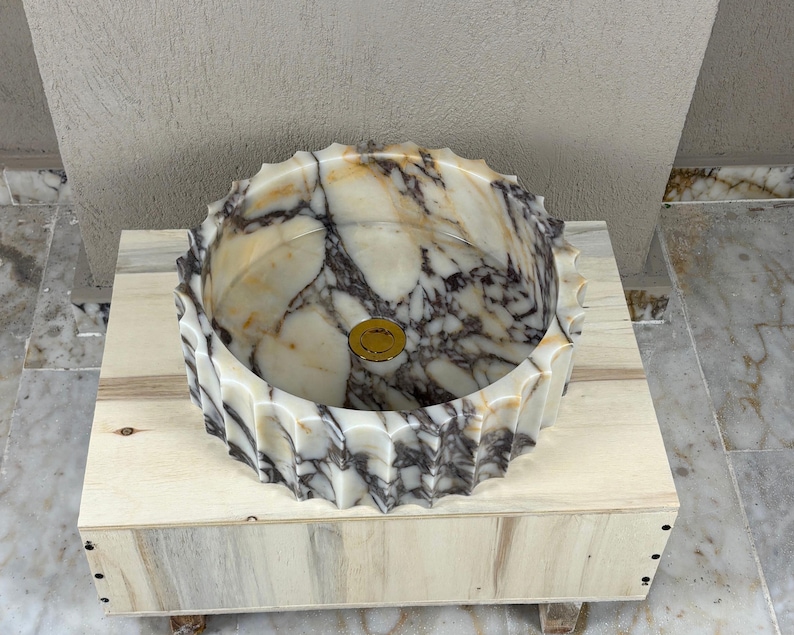 May include: A marble vessel sink with a scalloped edge. The sink features a cream and brown marbled pattern with a gold-colored drain cover. It is set on a light-colored wooden crate.
