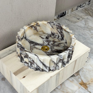 May include: A decorative marble sink with a unique, scalloped edge design. The sink features a blend of white, beige, and dark gray veining, and a gold-colored drain. It is set on a light-colored wooden base.