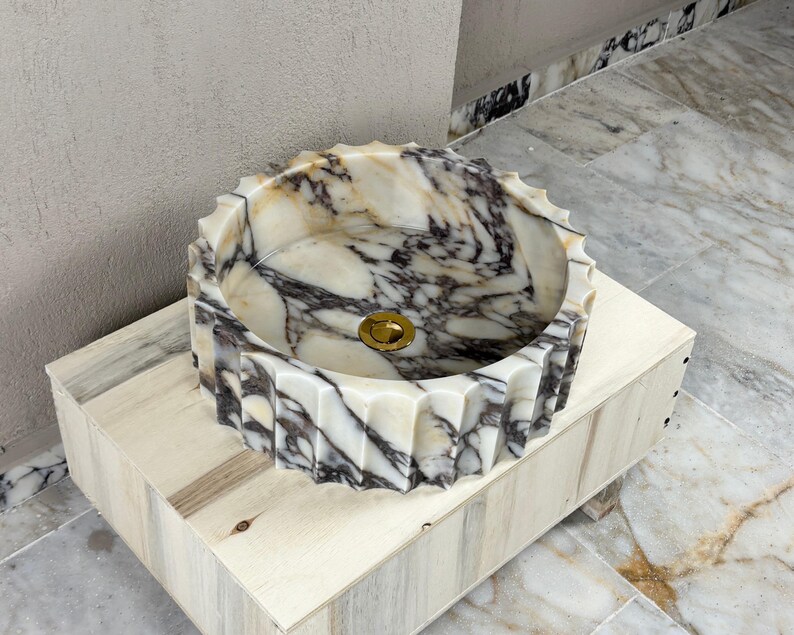May include: A decorative marble sink with a unique, scalloped edge design. The sink features a blend of white, beige, and dark gray veining, and a gold-colored drain. It is set on a light-colored wooden base.
