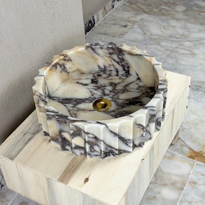 May include: A round marble sink with a scalloped edge. The sink features a white and gray veined pattern and a gold-colored drain. It is set on a light-colored wooden base.