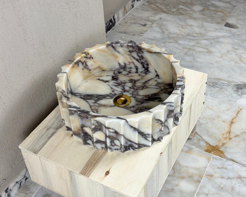 May include: A round marble sink with a scalloped edge. The sink features a white and gray veined pattern and a gold-colored drain. It is set on a light-colored wooden base.