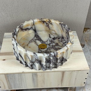 May include: A decorative marble sink with a unique, fluted design. The sink features a mix of white, beige, and dark gray veining. A gold-colored drain cover is visible in the center of the sink. The sink is displayed on a wooden base.