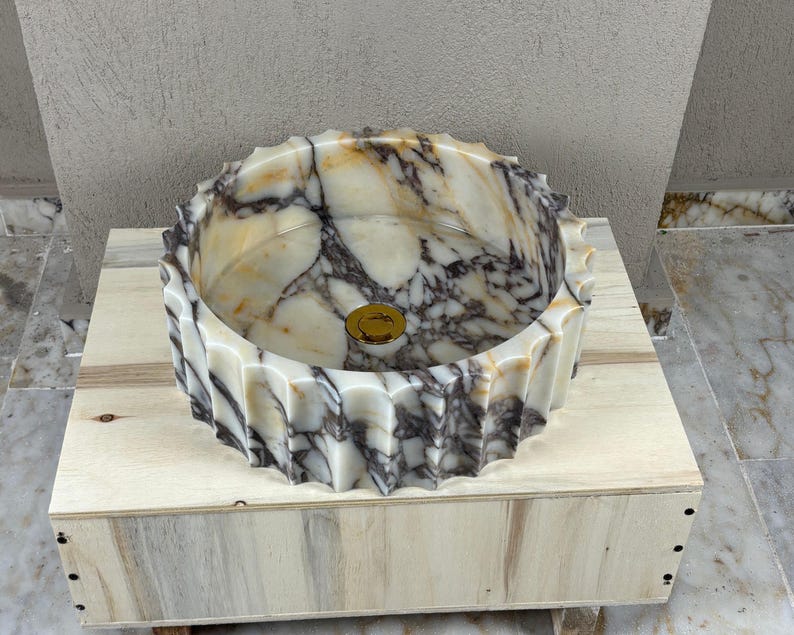 May include: A decorative marble sink with a unique, fluted design. The sink features a mix of white, beige, and dark gray veining. A gold-colored drain cover is visible in the center of the sink. The sink is displayed on a wooden base.